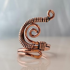 May include: A handcrafted copper wire ring with a unique, swirling design. The ring features a spiral and coiled wire elements, creating an artistic and eye-catching piece of jewellery. The ring is adjustable and has a polished finish.