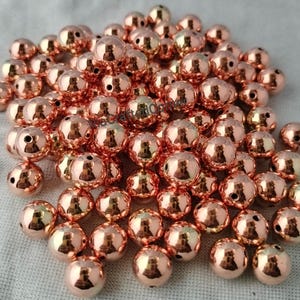 Könnte beinhalten: Eine große Ansammlung kleiner, kugelförmiger Perlen mit einer polierten, reflektierenden roségoldenen Metallic-Oberfläche. Jede Perle verfügt über ein einzelnes, mittiges Loch zum Auffädeln. Die Perlen liegen in einem losen, überlappenden Haufen und zeigen ihre glatte, glänzende Oberfläche sowie ihre gleichmäßige, runde Form.