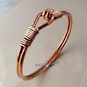 May include: A copper-colored bangle bracelet with a hook and eye closure. The bracelet features wrapped wire detailing at the closure and on one side. The overall design is simple and elegant, with a polished finish.