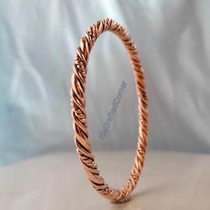 May include: A copper-colored, twisted metal bangle bracelet. The bracelet has a braided design and is displayed against a light blue background. The RiaCraftedCorner watermark is visible on the right side of the image.