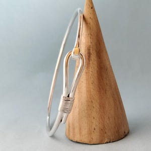 May include: A silver-tone bangle bracelet with a unique hook closure. The bracelet features a gold accent at the closure and a wrapped wire detail. The jewelry is displayed on a wooden cone against a light blue background.
