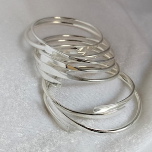 May include: A stack of silver-colored bangles. The bangles are thin, open-ended, and have a smooth, reflective surface. The bracelets are arranged in a loose pile, with some overlapping. The overall aesthetic is minimalist and modern.