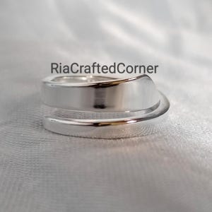 Könnte beinhalten: Nahaufnahme eines silbernen Rings mit löffelartigem Design. Der Ring umschließt den Finger mit einer glatten, polierten Oberfläche. Der Text "RiaCraftedCorner" ist auf der Oberseite des Rings zu sehen.