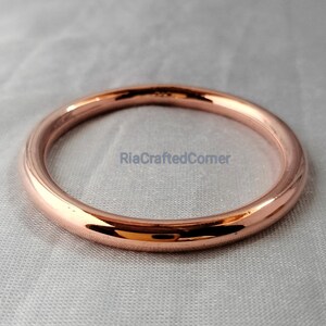 May include: A rose gold bangle bracelet. The circular bracelet is smooth and shiny, reflecting light. The bracelet is displayed on a light gray surface. The text "RiaCraftedCorner" is visible on the surface.