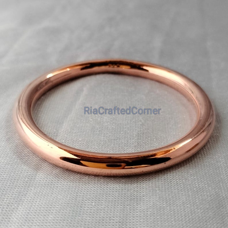 May include: A rose gold bangle bracelet. The circular bracelet is smooth and shiny, reflecting light. The bracelet is displayed on a light gray surface. The text "RiaCraftedCorner" is visible on the surface.