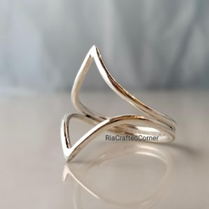 Może przedstawiać: Srebrny pierścionek o unikalnym, otwartym designie. Pierścionek posiada dwa elementy w kształcie litery V, które zachodzą na siebie, tworząc przyciągający wzrok geometryczny wzór. Pierścionek wykonany jest z błyszczącego, odbijającego światło metalu, a design jest minimalistyczny i nowoczesny.