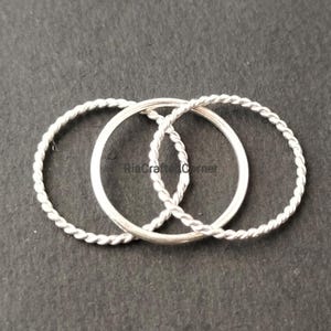 May include: Three silver rings are intertwined on a dark surface. Two rings have a twisted rope design, while the third is a smooth band. The rings are likely made of sterling silver. The text "RiaCraftedCorner" is visible.