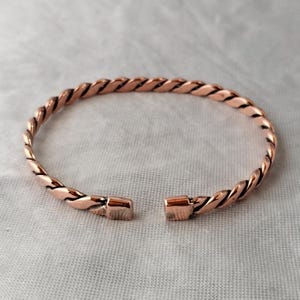 May include: A copper-colored bracelet with a twisted rope design. The open cuff bracelet has a rectangular clasp on each end. The jewelry is displayed on a light gray surface.