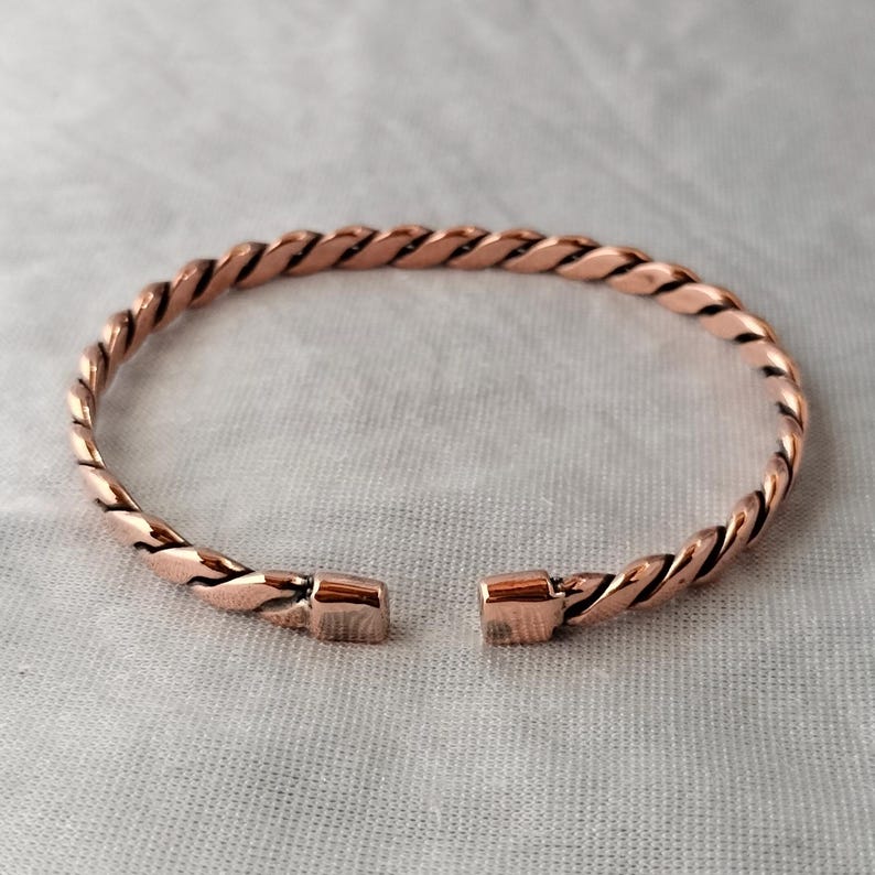 May include: A copper-colored bracelet with a twisted rope design. The open cuff bracelet has a rectangular clasp on each end. The jewelry is displayed on a light gray surface.