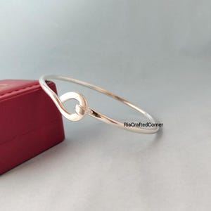 May include: A silver-toned bangle bracelet with a knot design. The bracelet is thin and delicate, with a smooth, polished finish. The knot detail is centered on the bracelet. The background is a light blue-gray.