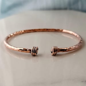 Op de afbeelding: Een koperkleurige open manchetarmband met een gehamerde textuur. De armband heeft een eenvoudig ontwerp met een gehamerde afwerking en decoratieve opgerolde uiteinden. Het open ontwerp maakt het gemakkelijk te dragen. De armband is een stijlvol accessoire.