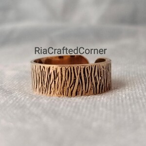 Puede incluir: Un anillo ajustable de color cobre con un patr&oacute;n texturizado similar a la corteza. El anillo tiene el texto "RiaCraftedCorner" en el fondo. El anillo est&aacute; abierto en la parte superior, lo que permite ajustar la talla. Es una joya.