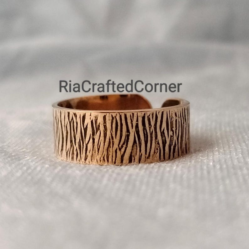 Puede incluir: Un anillo ajustable de color cobre con un patr&oacute;n texturizado similar a la corteza. El anillo tiene el texto "RiaCraftedCorner" en el fondo. El anillo est&aacute; abierto en la parte superior, lo que permite ajustar la talla. Es una joya.