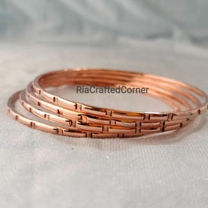 Pode incluir: Um conjunto de quatro pulseiras de cor cobre com um padrão repetitivo de entalhes retangulares na superfície externa. O texto RiaCraftedCorner é visível no centro das pulseiras de metal empilhadas, que possuem um acabamento polido e reflexivo.