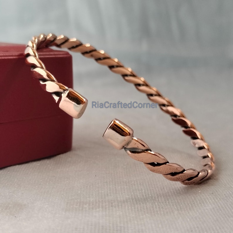 May include: A copper-colored, open cuff bracelet with a twisted rope design. The bracelet is made of metal and has a polished finish. The bracelet is shown on a light-colored surface with a red box in the background. The text "RiaCraftedCorner" is visible.