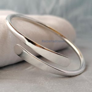 Puede incluir: Un brazalete de puño plateado pulido con un diseño minimalista. El brazalete tiene un acabado liso y una forma curvada, con extremos aplanados. Se muestra sobre una superficie clara y texturizada. El diseño del brazalete es simple y elegante.