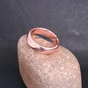 Può includere: Un anello in metallo lucido color oro rosa con una fascia liscia e arrotondata. L'anello presenta una superficie sottilmente riflettente ed è inciso con il testo RiaCraftedCorner sulla fascia interna, mostrando un design circolare pulito.