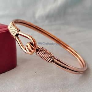 May include: A copper-colored bangle bracelet with a hook closure. The bracelet features a wrapped wire design at the hook and on the opposite side. The text "RiaCraftedCorner" is visible on the image.