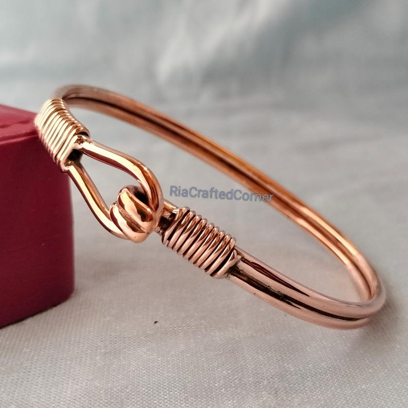 May include: A copper-colored bangle bracelet with a hook closure. The bracelet features a wrapped wire design at the hook and on the opposite side. The text "RiaCraftedCorner" is visible on the image.