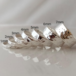 May include: A collection of silver rings of varying widths, from 1mm to 7mm. Each ring has a hammered texture, creating a unique, reflective surface. The rings are arranged in a diagonal line, showcasing their different sizes.