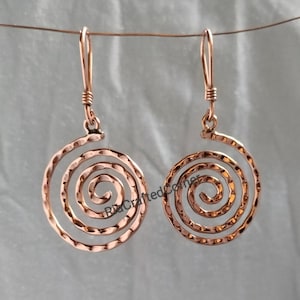 May include: A pair of copper-colored metal earrings featuring a circular spiral design with a hammered texture. Each earring hangs from a simple hook wire. The text RiaCraftedCorner is visible across the center of the spiral earring design.