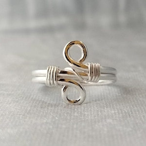 Puede incluir: Un anillo plateado con un diseño único. El anillo presenta una doble banda y está adornado con una forma abstracta envuelta en alambre con detalles dorados. El anillo es probablemente de plata de ley y es adecuado para el uso diario.