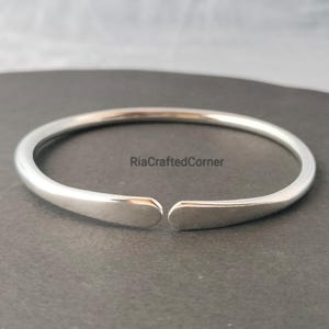 Peut inclure: Un bracelet manchette ouvert de couleur argentée. Le bracelet a une finition lisse et polie et un design minimaliste. Les extrémités du bracelet sont légèrement aplaties. Le texte "RiaCraftedCorner" est visible.