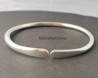 Könnte beinhalten: Ein silberfarbenes offenes Armreif-Armband. Das Armband hat eine glatte, polierte Oberfläche und ein minimalistisches Design. Die Enden des Armbands sind leicht abgeflacht. Der Text "RiaCraftedCorner" ist sichtbar.