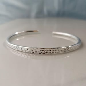 May include: A silver cuff bracelet with a hammered texture. The open-ended bracelet has a polished finish and reflects light. The bracelet is a simple, elegant piece of jewelry.