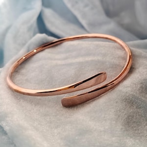 Peut inclure: Un bracelet manchette en or rose avec un design ouvert. Le bracelet a une finition lisse et polie et un style simple et élégant. Le bracelet est présenté sur un tissu bleu clair.
