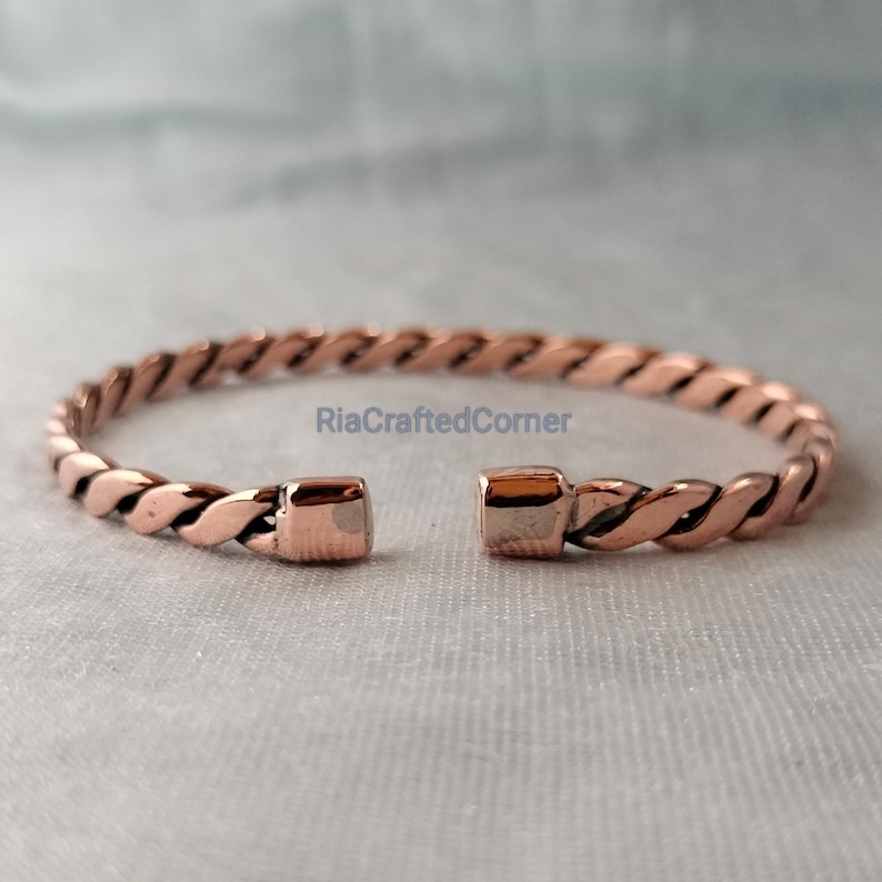 May include: A copper-colored, open cuff bracelet with a twisted rope design. The bracelet has a square clasp on each end. The text "RiaCraftedCorner" is visible on the bracelet.