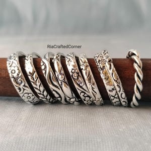 Puede incluir: Una colección de ocho anillos de metal plateado dispuestos sobre una varilla de madera oscura. Los anillos presentan varios patrones en relieve, incluyendo motivos florales, diseños de hojas, líneas onduladas y una textura de cuerda trenzada. El texto RiaCraftedCorner es visible.