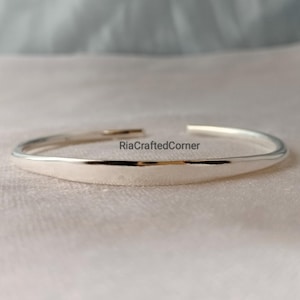 Könnte beinhalten: Ein silberfarbenes, offenes Armband. Das Armband hat eine glatte, polierte Oberfläche und ein leicht abgeflachtes Design. Der Text "RiaCraftedCorner" ist auf dem Armband sichtbar. Das Armband wird auf einer hellen Oberfläche präsentiert.