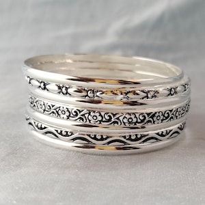 May include: A stack of silver bangles with detailed patterns. The bangles have floral, beaded, and wavy designs. The jewellery is displayed on a light-coloured surface.