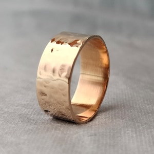 Könnte beinhalten: Ein gehammerter Goldring mit breitem Band. Der Ring hat eine strukturierte Oberfläche mit kleinen Vertiefungen, die ihm ein rustikales Aussehen verleihen. Der Ring besteht aus warmem, goldenem Metall und wird vor einem neutralen Hintergrund präsentiert.