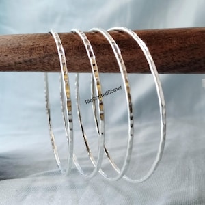 May include: A set of five silver bangles with a hammered texture, displayed on a wooden dowel against a light blue background. The bangles have a simple, elegant design. The text "RiaCraftedCorner" is visible on the dowel.
