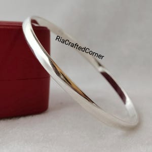 May include: A polished silver bangle with a smooth, rounded design. The bracelet is shown on a white surface beside a red box. The text "RiaCraftedCorner" is visible on the image.