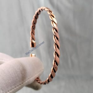 May include: A copper-colored, twisted-rope style bracelet. The open cuff design features a simple clasp. The bracelet is shown against a soft, neutral background. The text "RiaCraftedCorner" is visible on the image.
