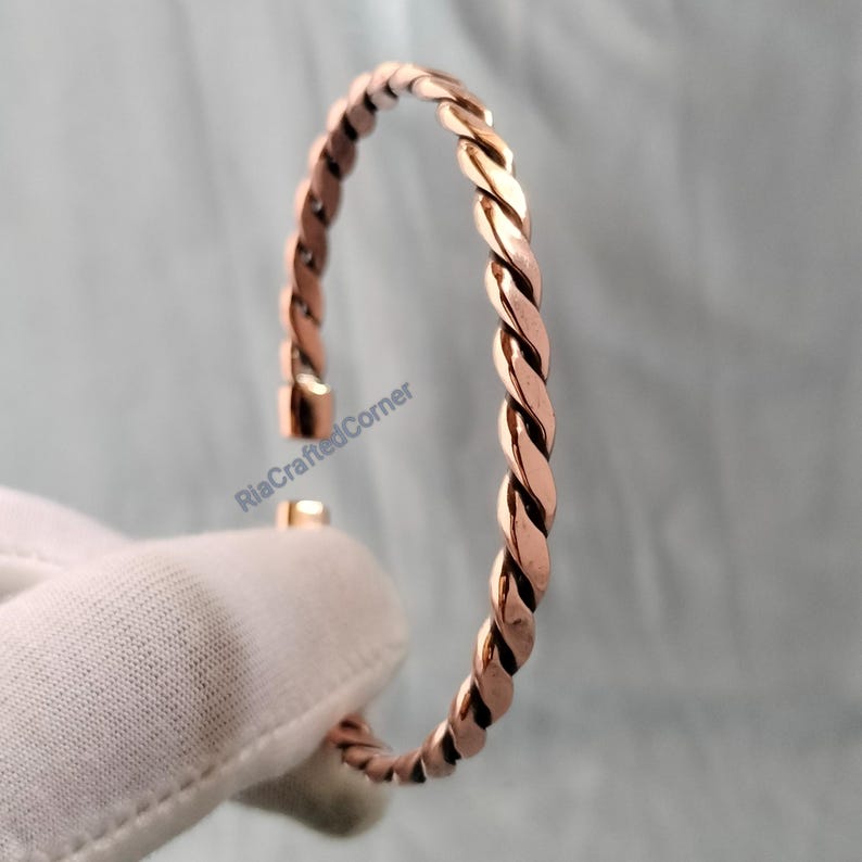 May include: A copper-colored, twisted-rope style bracelet. The open cuff design features a simple clasp. The bracelet is shown against a soft, neutral background. The text "RiaCraftedCorner" is visible on the image.