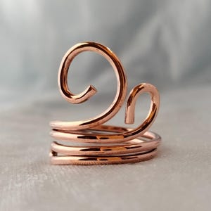 Puede incluir: Un anillo de color cobre con un diseño único. El anillo presenta espirales y acentos curvos, con múltiples bandas que se enrollan alrededor del dedo. El metal tiene un acabado pulido, que refleja la luz.