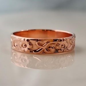May include: A rose gold ring with a floral pattern. The ring has a continuous design of flowers and swirling leaves carved around the band. The ring is highly polished, reflecting light and creating a warm appearance.