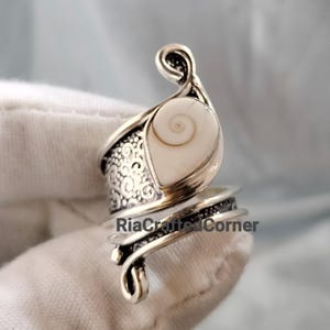 Puede incluir: Un anillo de plata con una concha de ojo de Shiva en forma de lágrima con un patrón de espiral natural. La banda está hecha de plata oxidada con intrincados grabados y detalles metálicos en espiral. El texto RiaCraftedCorner es visible.