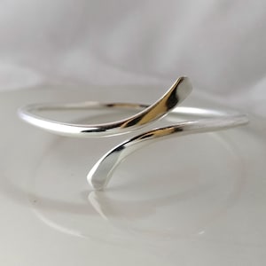 May include: A silver cuff bracelet with a minimalist, open design. The bracelet has a smooth, polished surface and a curved shape. The ends of the bracelet curve upwards, creating a stylish and elegant look. The bracelet is made of a shiny metal.