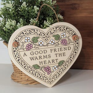 May include: Heart-shaped ceramic plaque with the text "A GOOD FRIEND WARMS THE HEART". The plaque is cream-colored with a decorative border and floral accents in pink, green, and white. It hangs from a natural twine cord.