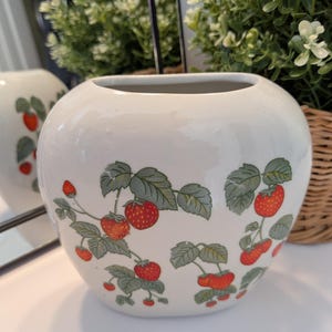 May include: A white ceramic vase with a round shape. The vase is decorated with a pattern of red strawberries and green leaves. The vase is likely used for decorative purposes.