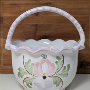 May include: White ceramic basket with a scalloped rim and a handle. The handle is white with pink polka dots. The basket is decorated with a pink and purple floral design and green leaves.