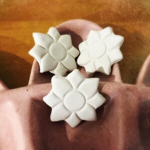 May include: Three white, matte-finish, flower-shaped objects with eight petals and a central circular disc. The items are arranged in a cluster, each featuring a smooth, uniform surface texture and a distinct, symmetrical floral design.