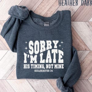 May include: Heather dark gray sweatshirt with the text "SORRY I'M LATE HIS TIMING, NOT MINE" in white. The text also includes "ECCLESIASTES 3:1". The sweatshirt is folded on a light brown and white wooden surface.