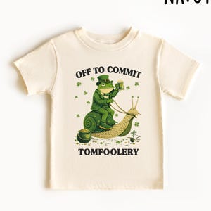 May include: Cream-colored t-shirt featuring a whimsical illustration. A green frog in a leprechaun outfit rides a snail, holding a beer. The text "OFF TO COMMIT TOMFOOLERY" is printed above and below the image, with shamrocks scattered around.