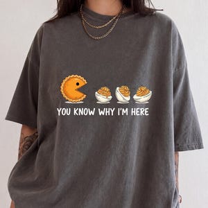 May include: A charcoal gray short-sleeve t-shirt featuring a graphic of a circular orange pastry with a mouth open, chasing three halved deviled eggs. Below the graphic, the white text reads, "YOU KNOW WHY I'M HERE" in a bold, sans-serif font.
